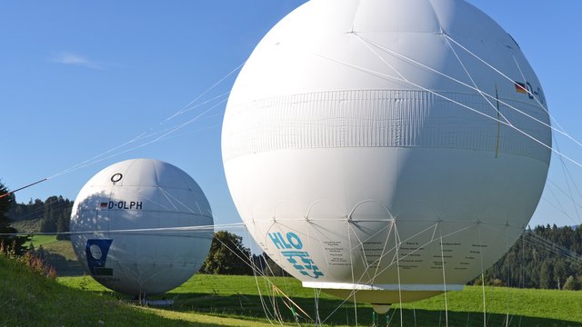 CL- und HLO-Ballon Zwei große Ballone stehen auf einem grünen Feld. Der vordere Ballon hat ein Logo mit der HLO. Der hintere, etwas kleinere Ballon trägt die Kennung D-OLPH. Im Hintergrund sind Bäume und blauer Himmel zu sehen.