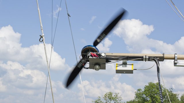Elektroantrieb des Airkules Ein Propeller befindet sich an einem Tragarm, vor einem Himmel mit Wolken. Der Propeller dreht sich und ist mit elektrischen Leitungen verbunden. Im Hintergrund sind Bäume und andere Rigging-Elemente zu sehen.